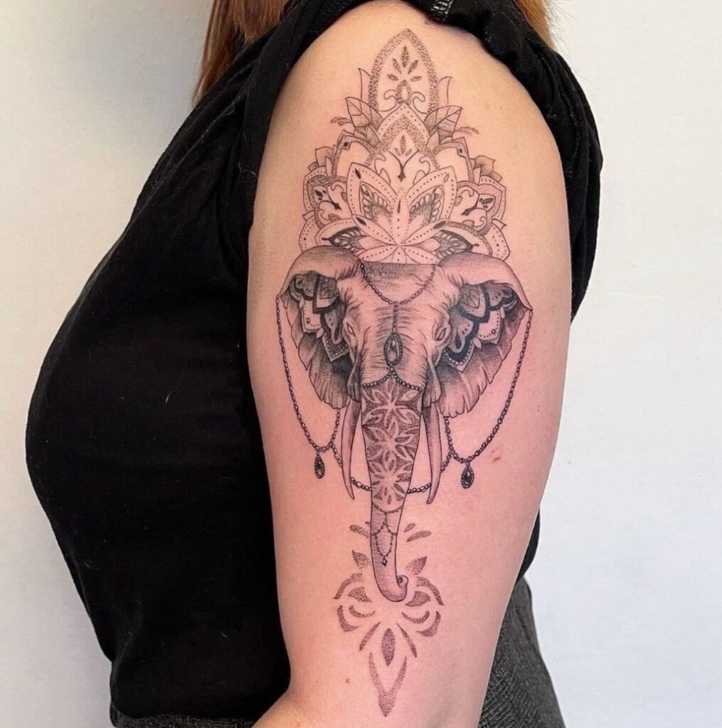Een verfijnde fineline tattoo op een bovenarm van een olifantenkop, rijkelijk versierd met ornamentele patronen, mandala-details en subtiele parelsnoeren. De tattoo is uitgevoerd in een combinatie van dun lijnwerk en zacht dotwork. Werk van Yifei (Herz Tattoo) tijdens de Sint-Niklaas Tattoo Expo.