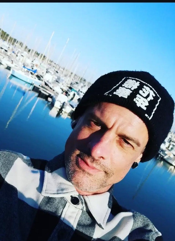 Een selfie van tattoo-artiest Olivier (olivier.barre.108) bij een jachthaven. Hij draagt een zwarte muts met Japanse tekens en een zwart-wit geruit hemd. De zonnige achtergrond toont talloze zeilboten tegen een helderblauwe lucht.