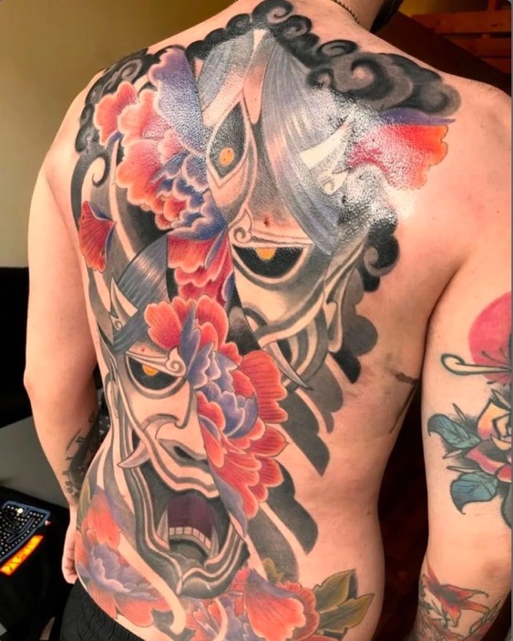 Een grootschalige, kleurrijke tattoo op de rug die Japanse elementen combineert. Het ontwerp toont een gedetailleerd Hannya-masker en grote florale patronen in rode, paarse en grijze tinten, omlijst door donkere wolkenpartijen. Werk van Olivier (olivier.barre.108) op de Sint-Niklaas Tattoo Expo.