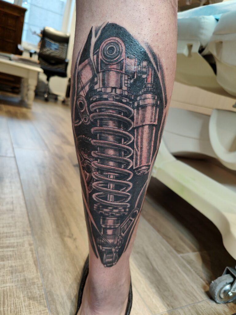 Een gedetailleerde biomechanische tattoo op een onderbeen, waarbij een realistische schokdemper en veer lijken te verschijnen onder de huid. De tattoo is uitgevoerd in black and grey met sterke contrasten. Gezet door Johnny van Johnny's Tattoos Hamme op de Sint-Niklaas Tattoo Expo.