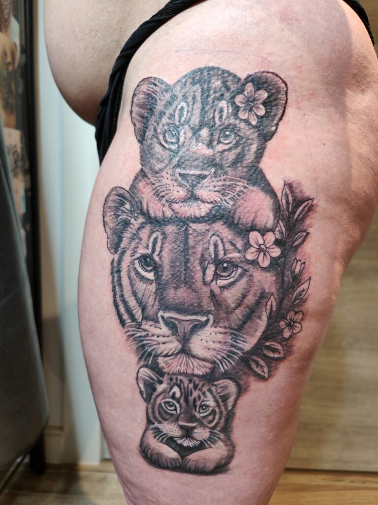 Een aandoenlijke black and grey tattoo op een bovenbeen van een leeuwin met haar twee welpen. De compositie is verticaal opgebouwd en versierd met subtiele botanische elementen aan de zijkant. Werk van Johnny (Johnny's Tattoos Hamme) op de Sint-Niklaas Tattoo Expo.