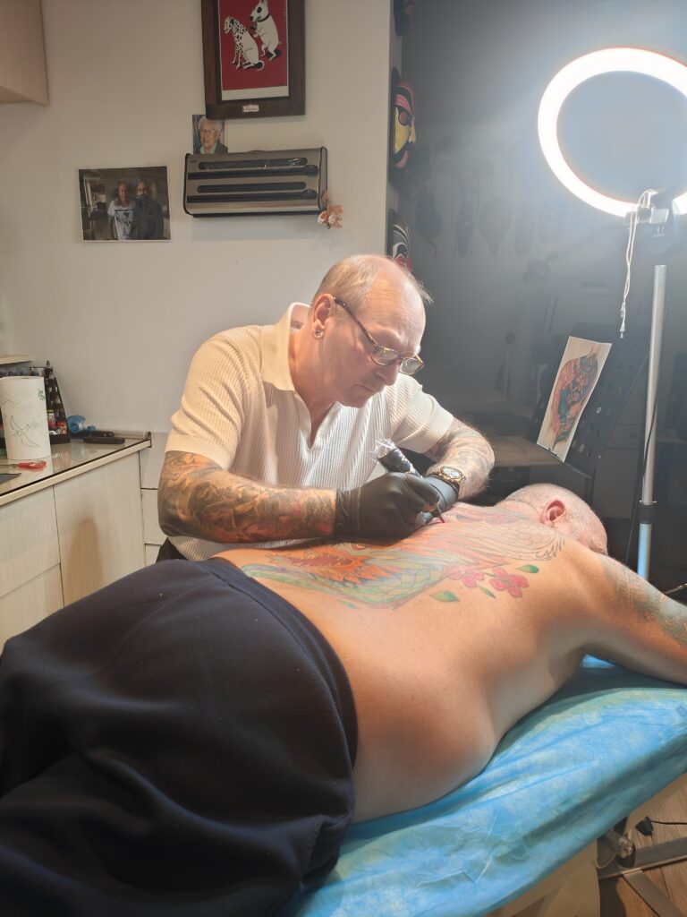 Een actiefoto van tattoo-artiest Johnny van Johnny's Tattoos Hamme tijdens de Sint-Niklaas Tattoo Expo. Hij draagt een witte polo en bril, en is geconcentreerd bezig met het inkleuren van een grootschalige, kleurrijke rugtattoo van een draak onder een professionele ringlamp.