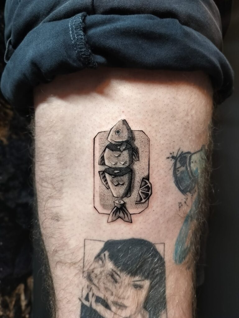 Een unieke, grafische tattoo van een in mootjes gesneden vis op een serveerplankje met een schijfje citroen. De tattoo is uitgevoerd in blackwork met fijn dotwork voor textuur. Werk van Nika (Unpunktual Tattoo) op de Sint-Niklaas Tattoo Expo.