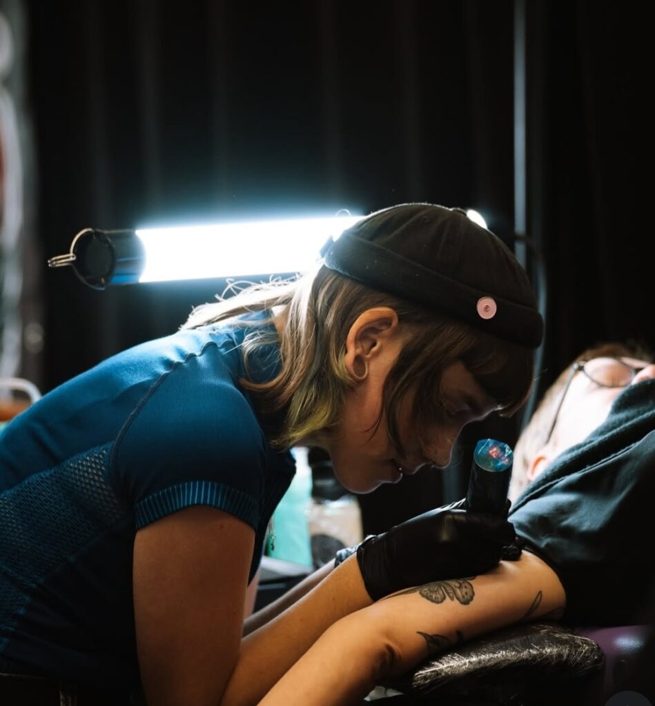 Sfeerbeeld van tattoo-artiest Nika (Unpunktual Tattoo) terwijl ze geconcentreerd een vlinder tatoeëert op de Sint-Niklaas Tattoo Expo. Ze draagt een zwarte muts en een blauw shirt, terwijl ze onder fel studiolicht met een draadloze tattoo-machine werkt.