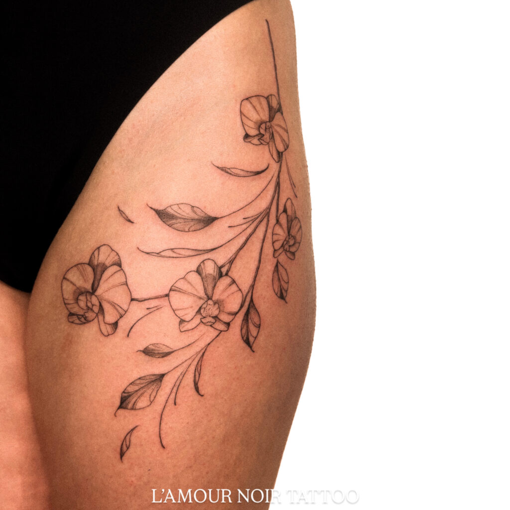 Een verfijnde fineline tattoo van een tak met orchideeën en bladeren op een bovenbeen. Het ontwerp kenmerkt zich door uiterst dunne lijnen en zachte schaduwwerking, wat zorgt voor een elegante en vrouwelijke uitstraling. Werk van Chelsy (L'amour Noir Tattoo) op de Sint-Niklaas Tattoo Expo.
