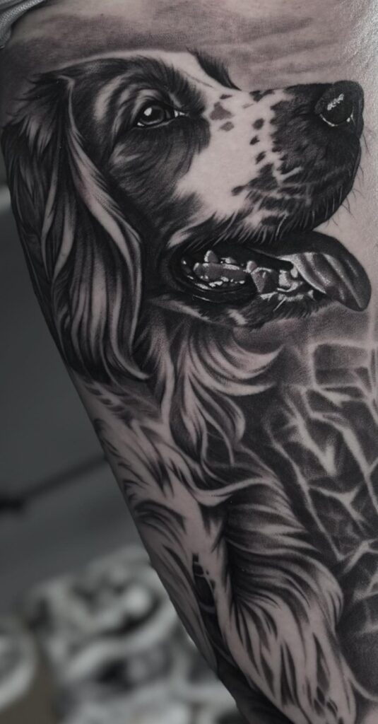 Een hyperrealistisch black and grey portret van een hond op een bovenarm. De tattoo toont de hond van opzij met de tong uit de bek, waarbij de glans in de ogen en de textuur van de zachte vacht met grote precisie zijn uitgewerkt. Gezet door Dante van Dante Ink op de Sint-Niklaas Tattoo Expo.