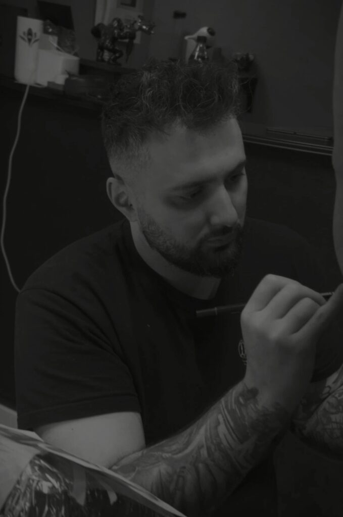 Een sfeervolle zwart-wit foto van tattoo-artiest Dante (Dante Ink) tijdens de Sint-Niklaas Tattoo Expo. Hij is geconcentreerd bezig met het voortekenen of perfectioneren van een ontwerp op de huid. Zijn eigen armen zijn voorzien van uitgebreide tatoeages.