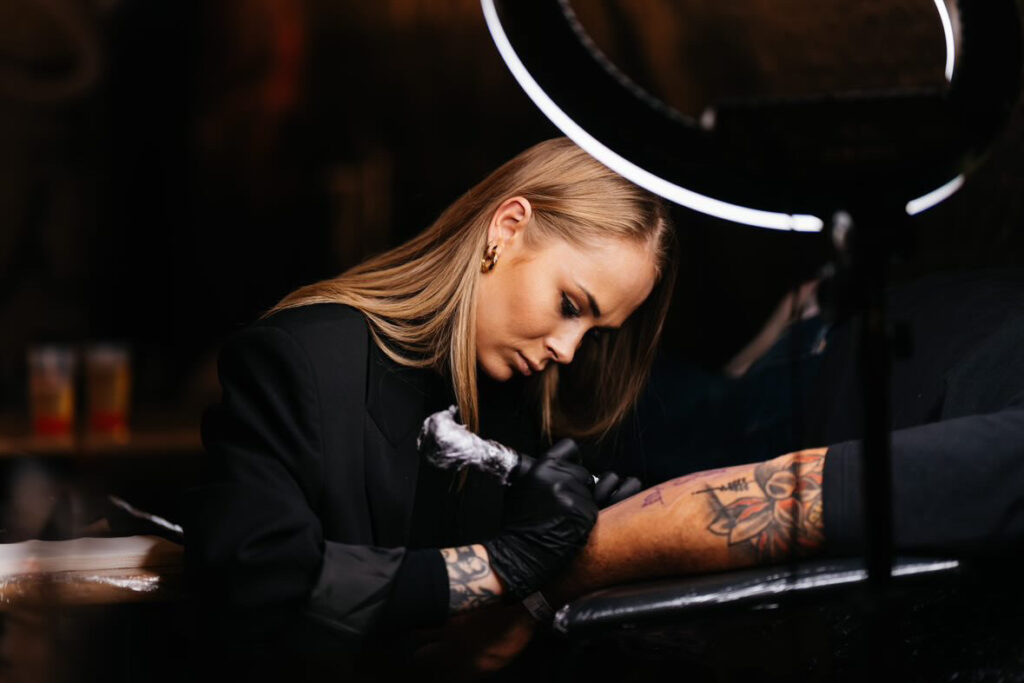 Een sfeervolle actiefoto van tattoo-artiest Chelsy (L'amour Noir Tattoo) tijdens de Sint-Niklaas Tattoo Expo. Ze is geconcentreerd bezig met het zetten van een kleurrijke tattoo op een arm onder professionele ringverlichting. Ze draagt een zwarte blazer en zwarte handschoenen.