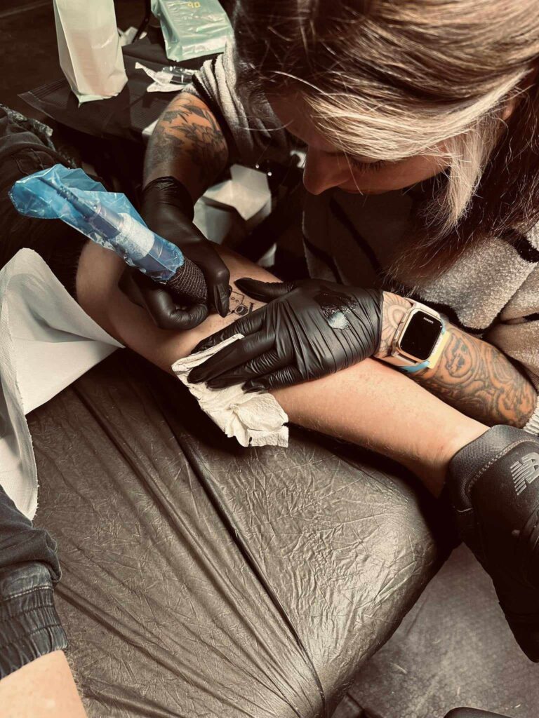 Een close-up van tattoo-artiest Ellen (Black Swan Tattoo) terwijl ze geconcentreerd een tatoeage zet op de Sint-Niklaas Tattoo Expo. Ze draagt zwarte handschoenen en werkt met een professionele tattoo-machine, terwijl haar eigen gedetailleerde arm-tattoos zichtbaar zijn.