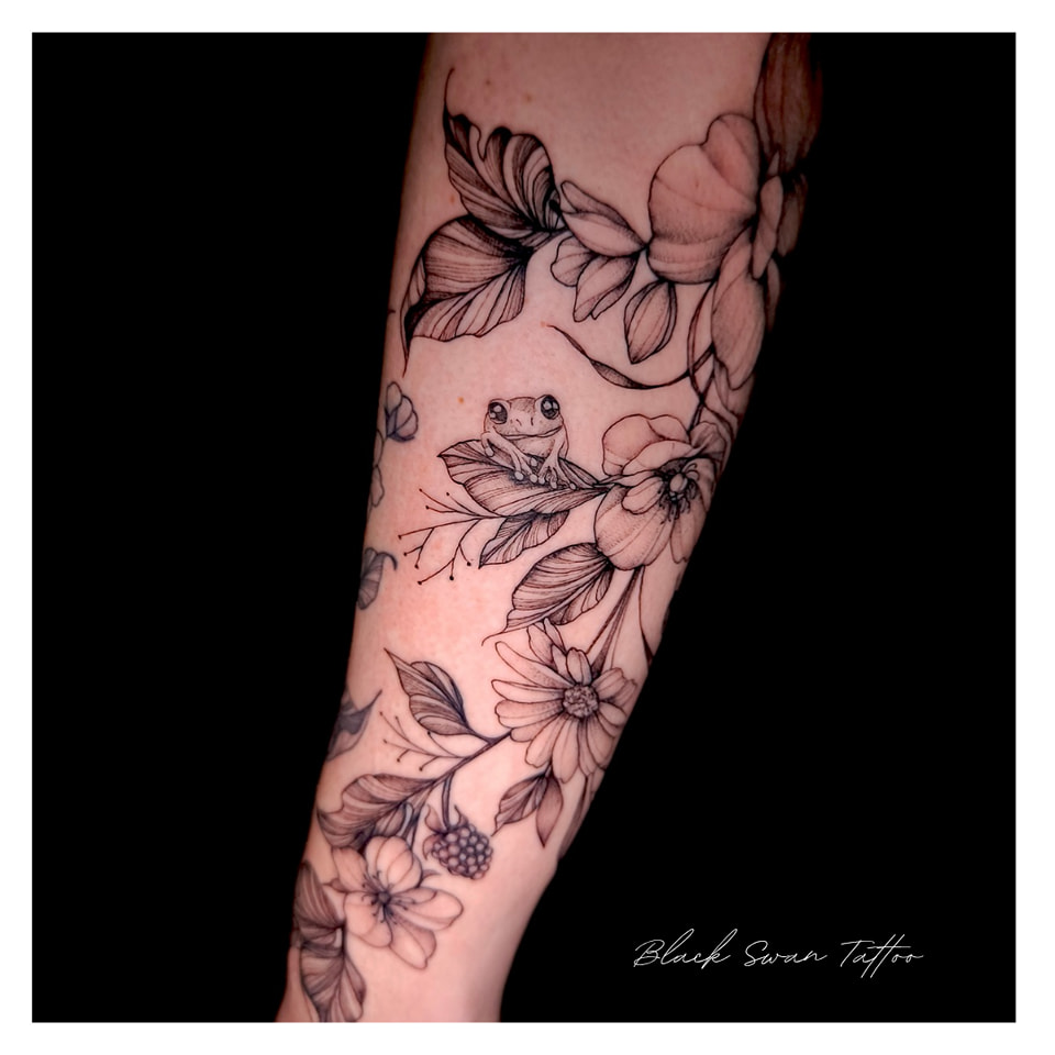 Een verfijnde fine-line tattoo op een onderarm met een schattig kikkertje dat op een blad tussen wilde bloemen en bramen zit. Het ontwerp is uitgevoerd in een lichte black and grey stijl met subtiele dotwork. Werk van Ellen (Black Swan Tattoo) op de Sint-Niklaas Tattoo Expo.