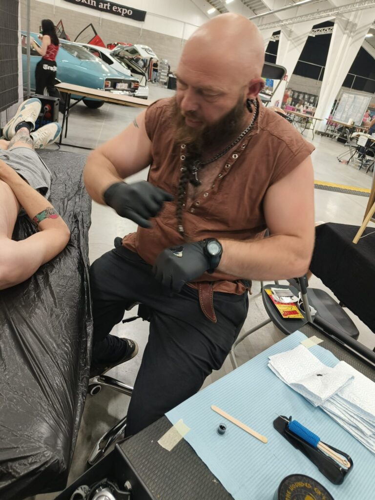Een actiefoto van tattoo-artiest Wim Geysen tijdens de Sint-Niklaas Tattoo Expo. Wim heeft een volle baard en draagt een bruine mouwloze top en zwarte handschoenen. Hij bereidt zijn werkplek voor om een klant te tatoeëren volgens traditionele methoden.