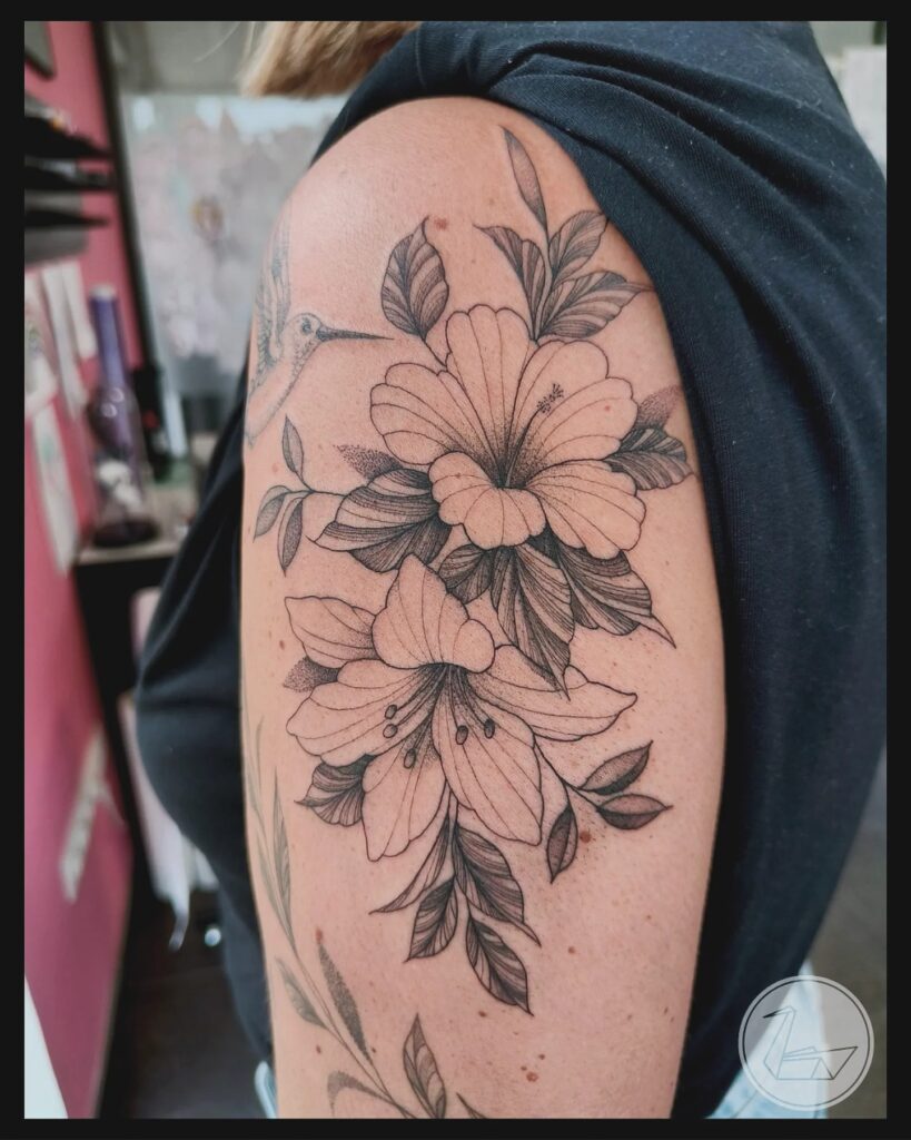 Gedetailleerde botanische tattoo op een schouder en bovenarm, bestaande uit hibiscus- en leliebloemen met fijn gearceerde bladeren. Op de achtergrond is een subtiele kolibrie zichtbaar. Gezet door Ellen van Black Swan Tattoo tijdens de Sint-Niklaas Tattoo Expo.