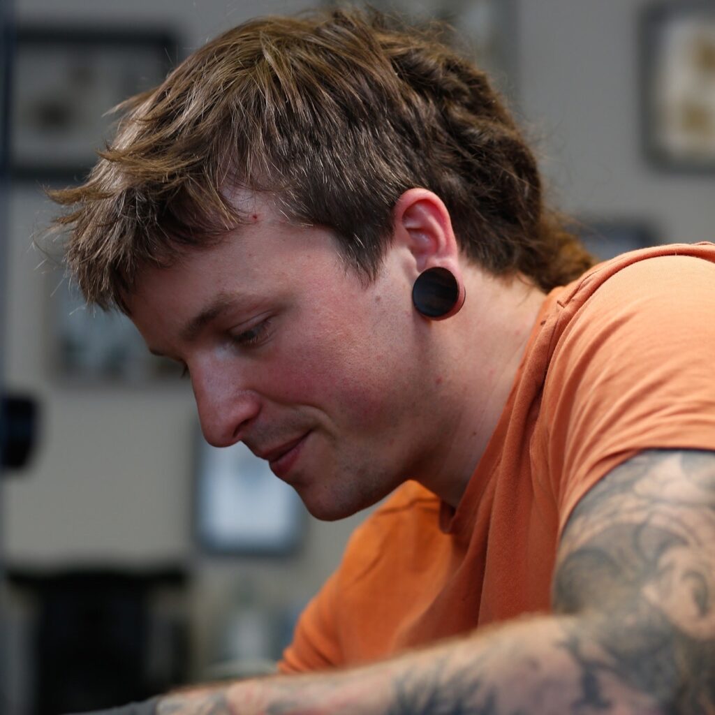 Een close-up portret van tattoo-artiest Simon (Sunrise Skin Art) aan het werk op de Sint-Niklaas Tattoo Expo. Hij draagt een oranje shirt en heeft zichtbare tattoos op zijn armen, geconcentreerd kijkend naar zijn werkstation.