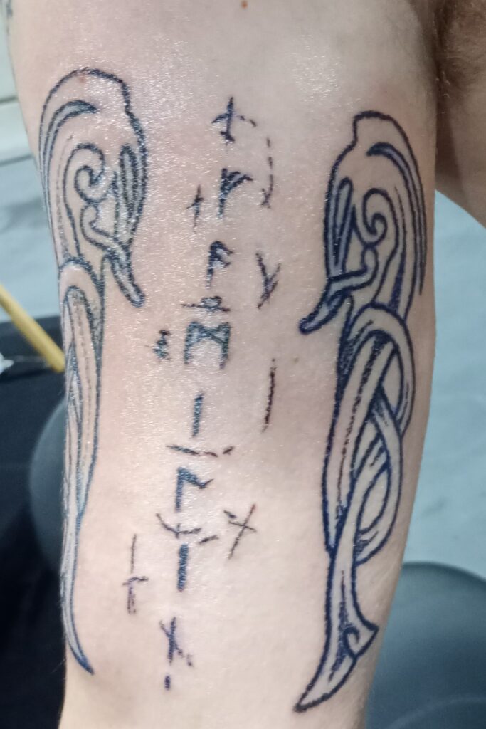 Een close-up van een tattoo in wording op een arm, bestaande uit een verticale rij Noorse runen geflankeerd door twee gestileerde vlechtwerkpatronen. Het ontwerp toont de authentieke, handgezette stijl van Wim Geysen op de Sint-Niklaas Tattoo Expo.