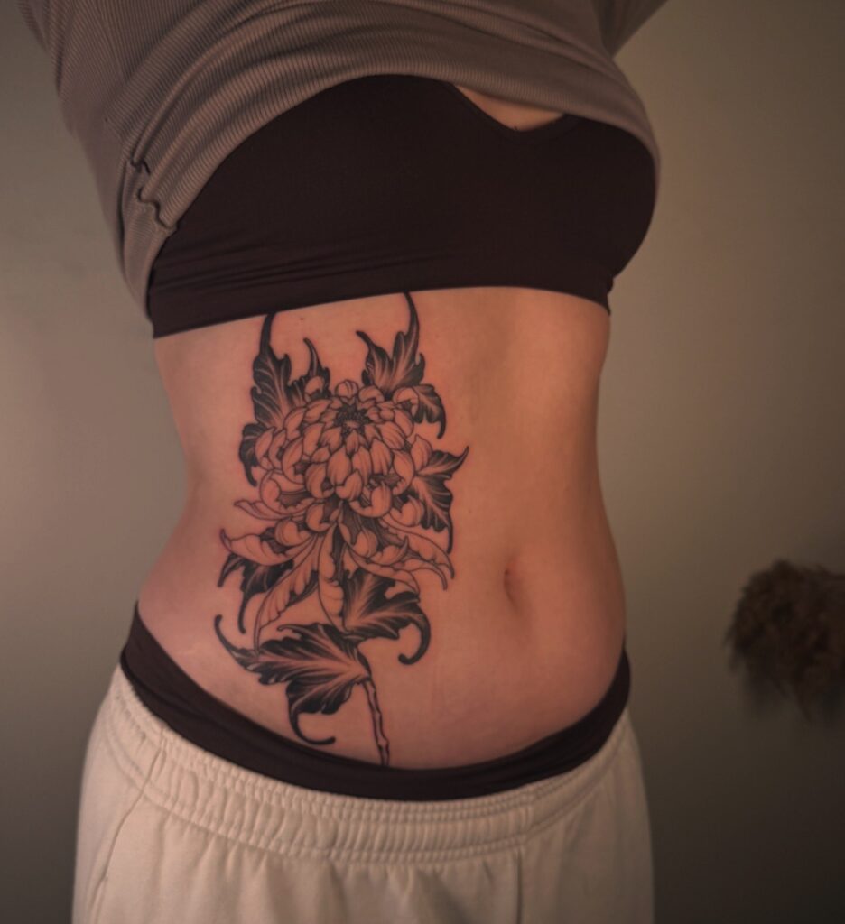 Een krachtige black and grey tattoo van een Japanse chrysant op de zij en ribben. Het ontwerp heeft diepe contrasten en dynamische bladeren die de vorm van het lichaam accentueren. Gezet door Simon van Sunrise Skin Art op de Sint-Niklaas Tattoo Expo."
