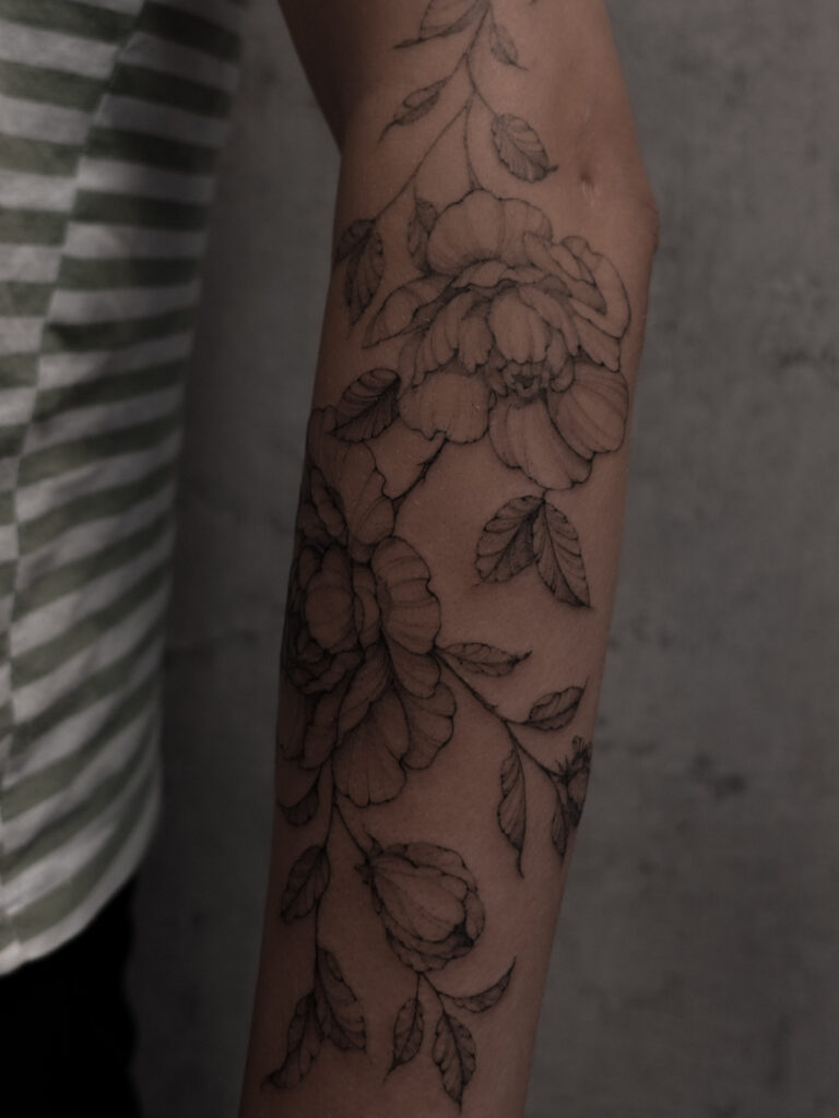 Een verfijnde fineline tattoo op de onderarm met gedetailleerde pioenrozen en bladeren. De tattoo loopt elegant over de arm met dunne lijnen en zachte dotwork-schaduwen. Gezet door Sasha van Sasha Hemelaer Tattoo op de Sint-Niklaas Tattoo Expo.