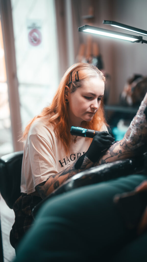 Een sfeervolle actiefoto van tattoo-artiest Jana (Jana Jacques Tattoo) op de Sint-Niklaas Tattoo Expo. Ze is geconcentreerd bezig met het tatoeëren van een arm onder een heldere studiolamp. Jana heeft oranjerood haar en draagt een beige T-shirt en zwarte handschoenen.