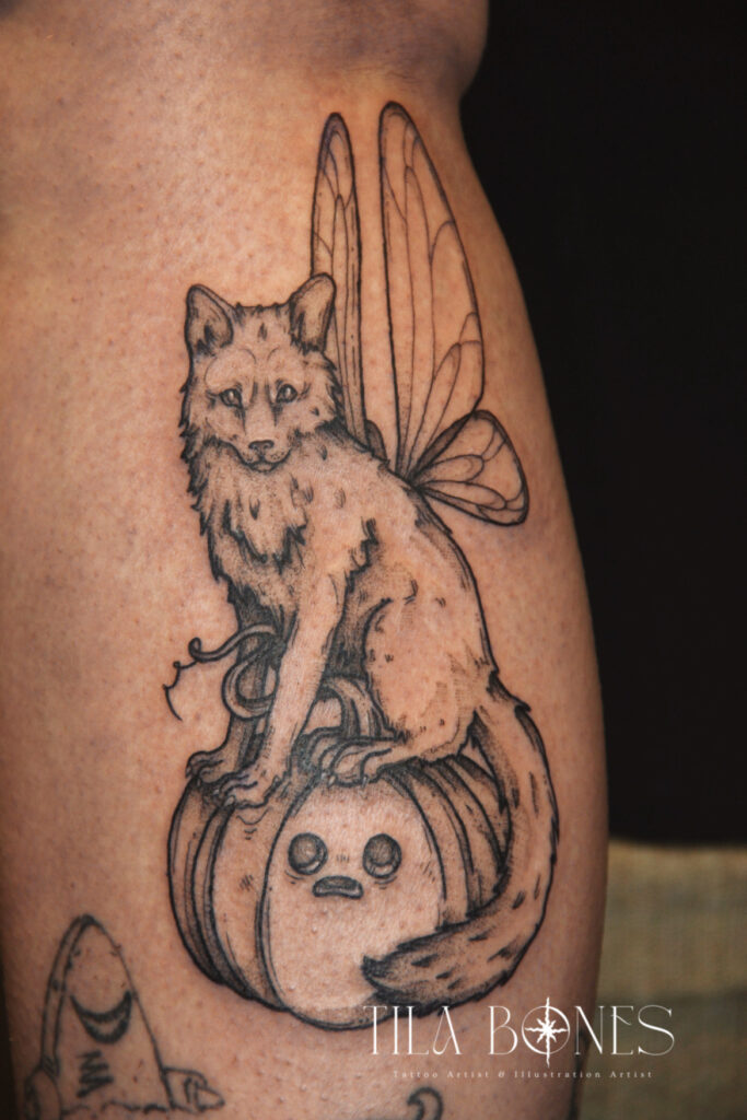 Een fantasierijke tattoo van een vos met vlindervleugels, zittend op een eigenzinnige pompoen met een gezichtje. De tattoo is uitgevoerd in een kenmerkende schetsmatige blackwork stijl. Gezet door Phoebe (Tila Bones) op de Sint-Niklaas Tattoo Expo.