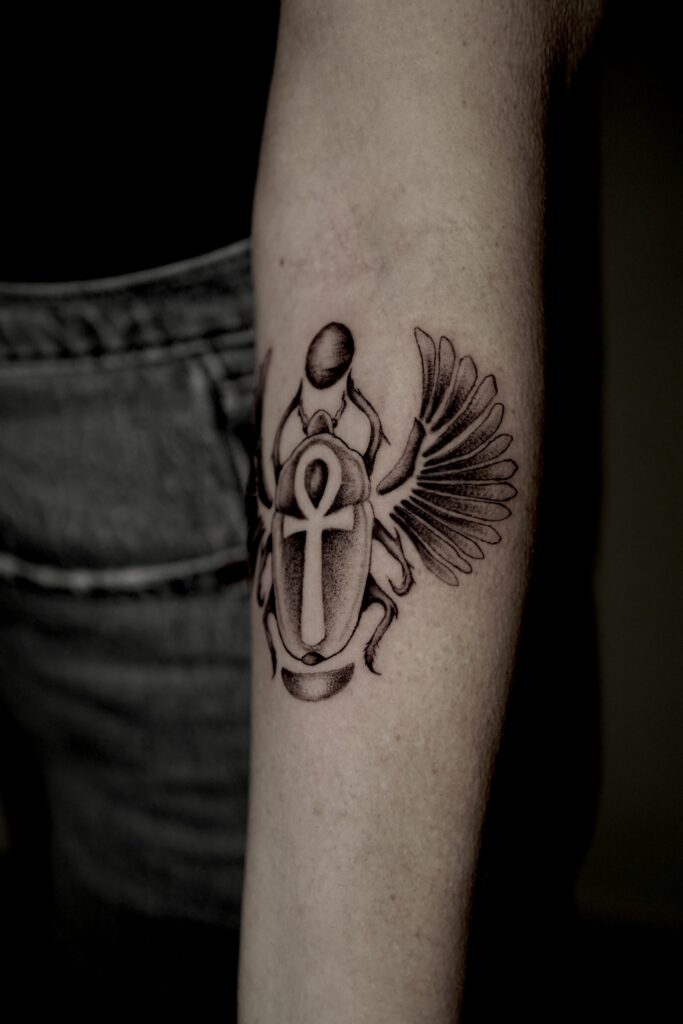 Een gedetailleerde fineline tattoo van een Egyptische scarabee op een onderarm. De scarabee houdt een zonneschijf vast en heeft een Ankh-symbool op zijn schild. Het ontwerp is uitgevoerd in black and grey met fijn schaduwwerk en gedetailleerde vleugels. Gezet door Naviia van Be Inkspired Tattoo op de Sint-Niklaas Tattoo Expo.