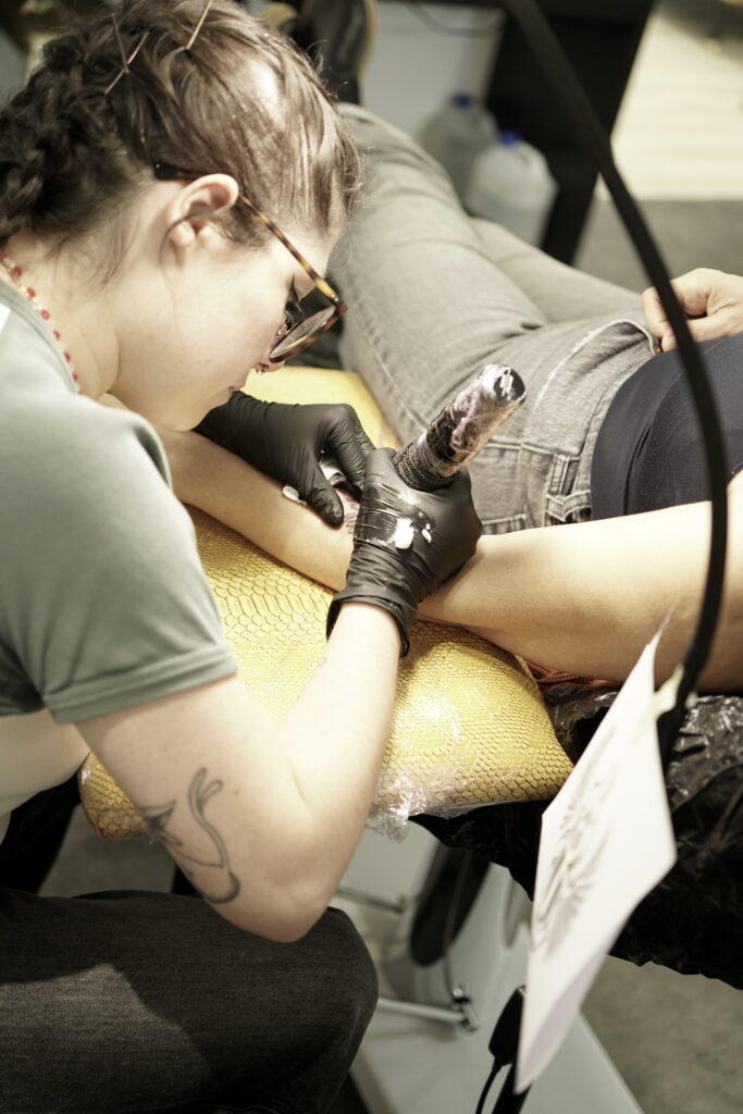 Een actiefoto van tattoo-artiest Naviia (Be Inkspired Tattoo) op de Sint-Niklaas Tattoo Expo. Ze draagt een bril en zwarte handschoenen terwijl ze geconcentreerd een tatoeage zet op de arm van een klant. Haar professionele houding en focus op detail zijn duidelijk zichtbaar.