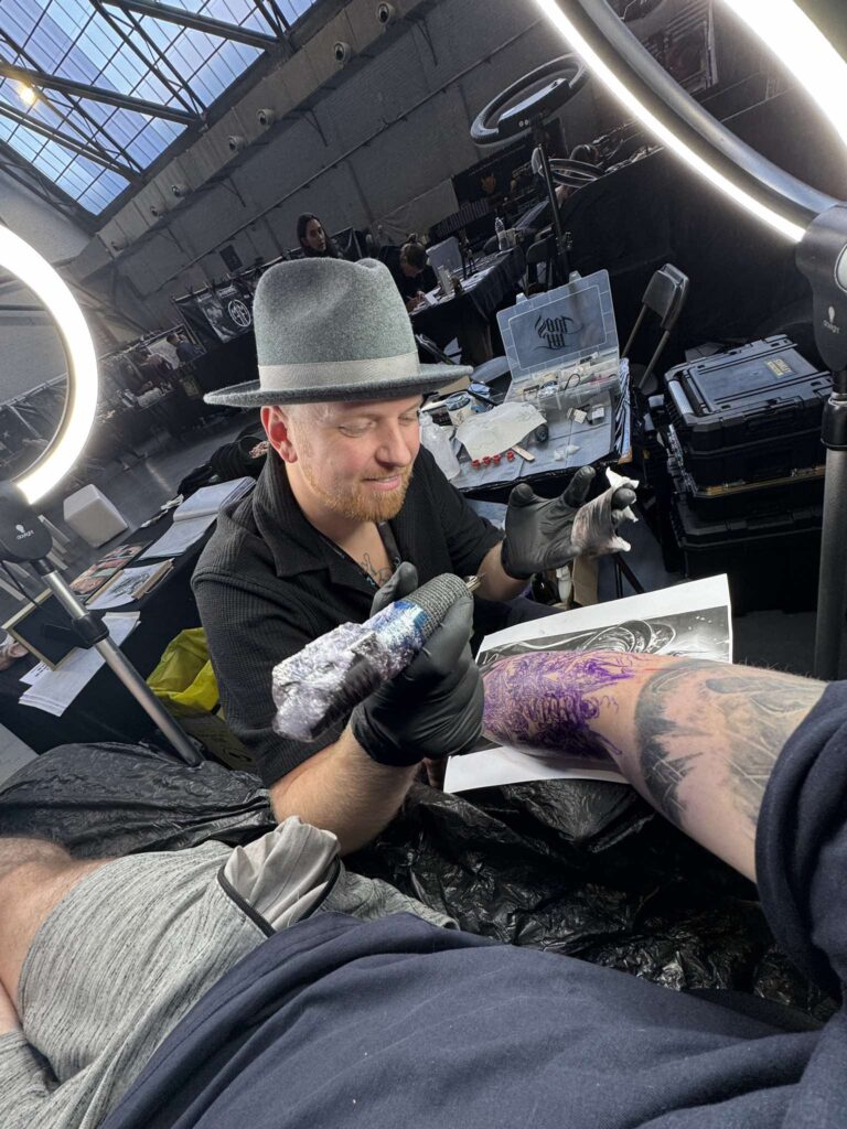 Een actiefoto van tattoo-artiest Michael (Martinez Tattoo) aan het werk op de Sint-Niklaas Tattoo Expo. Hij draagt een grijze hoed en zwarte handschoenen terwijl hij met precisie een paarse stencil op een arm tatoeëert onder professionele ringverlichting.
