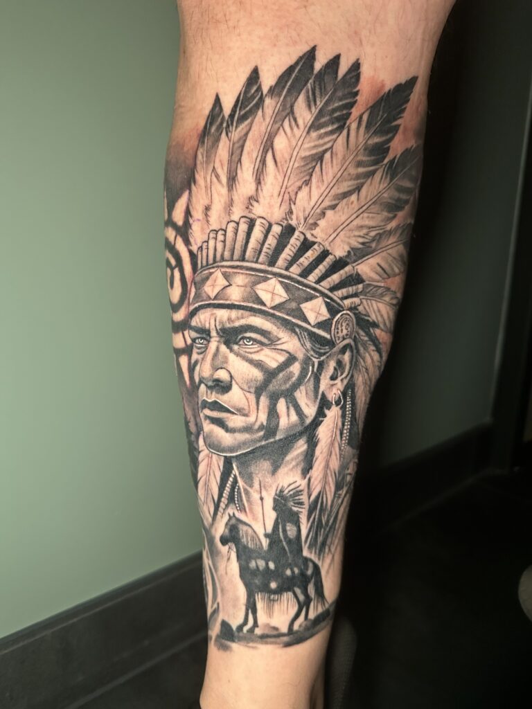 Gedetailleerd black and grey tattoo-portret van een inheemse Amerikaanse man met een indrukwekkende verentooi op een onderbeen. Onderaan de tattoo is een silhouet van een ruiter te paard zichtbaar. Vakmanschap van Michael (Martinez Tattoo) op de Sint-Niklaas Tattoo Expo.
