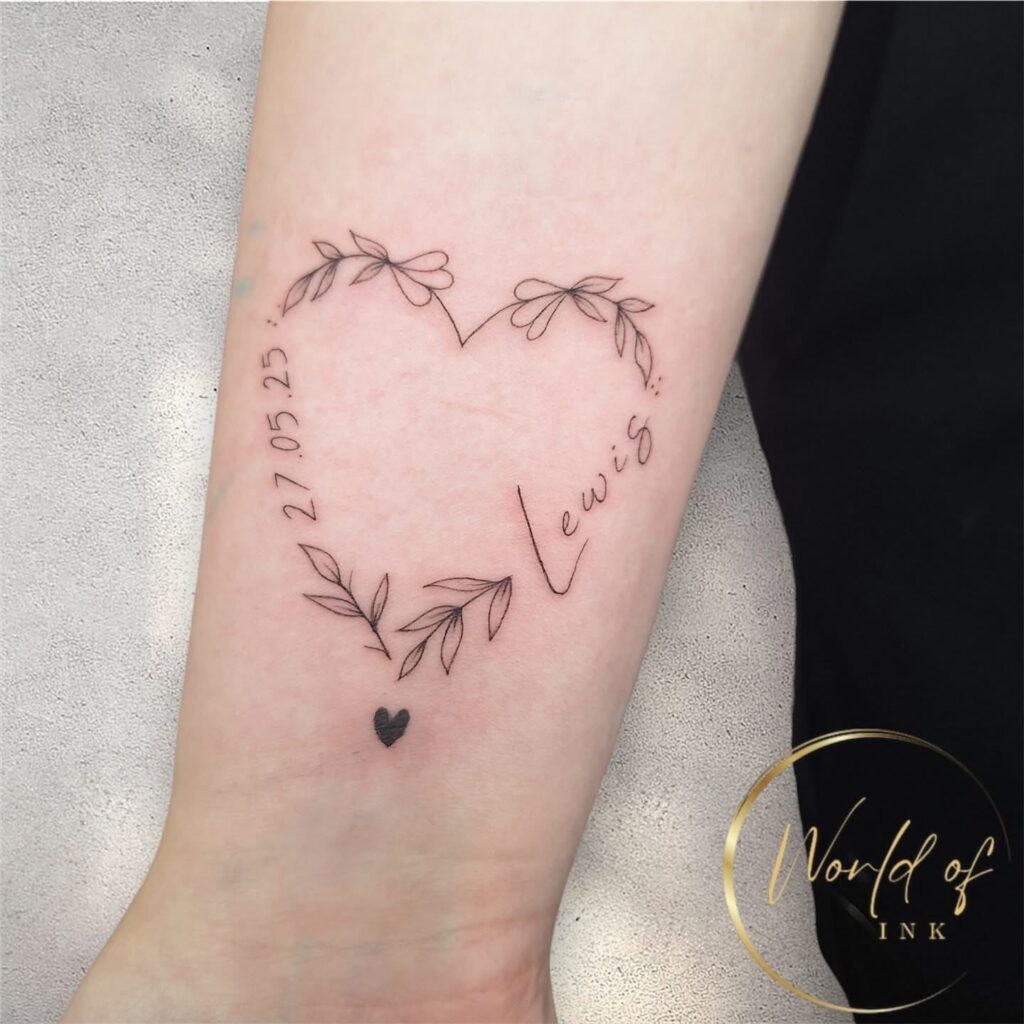 Een verfijnde fineline tattoo op een onderarm in de vorm van een hart, samengesteld uit sierlijke takjes met blaadjes. In het hart staan de naam Lewis en de datum 27.05.25 in een elegant handschrift. Onderaan het hart is een klein, zwart ingekleurd hartje te zien. Werk van Angelika (Go Ink Tattoos) op de Sint-Niklaas Tattoo Expo.