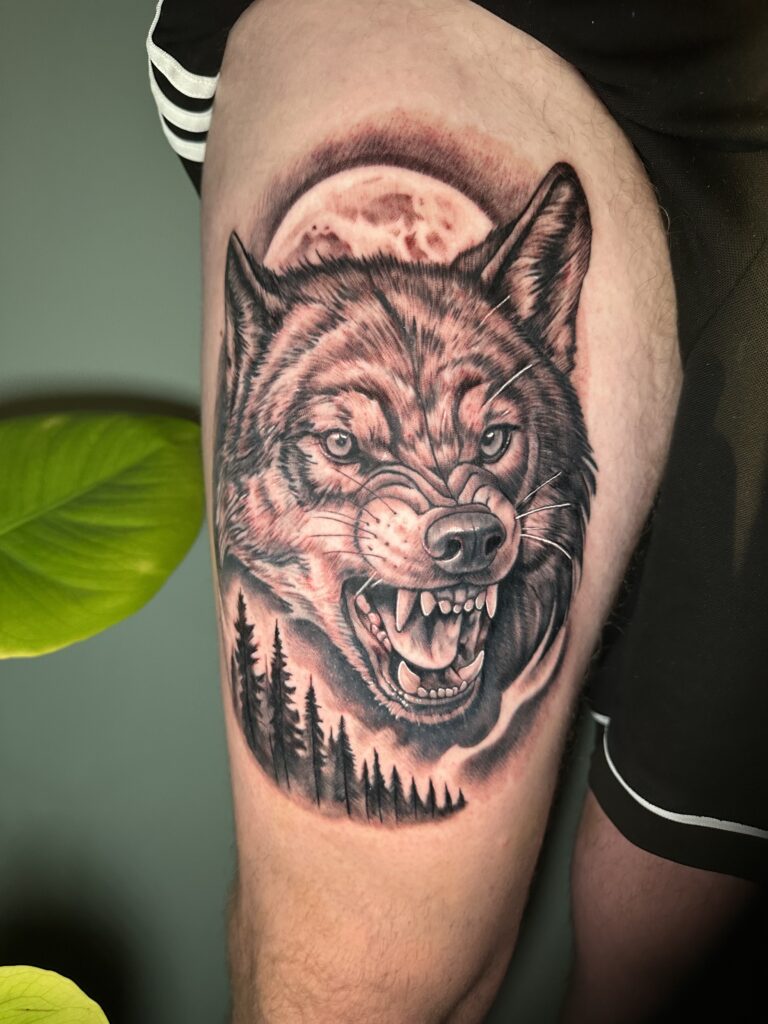 Een krachtige, realistische black and grey tattoo van een grommende wolf op een bovenbeen. De compositie bevat een volle maan op de achtergrond en silhouetten van dennenbomen aan de onderzijde. Gezet door Michael van Martinez Tattoo op de Sint-Niklaas Tattoo Expo.