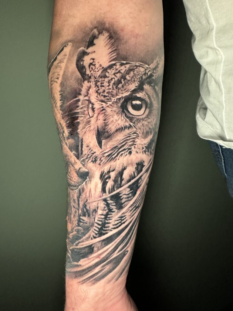 Een close-up van een zeer gedetailleerde uil-tattoo op een onderarm in realistische black and grey stijl. De focus ligt op de doordringende blik en de fijne textuur van de veren. Werk van Michael van Martinez Tattoo tijdens de Sint-Niklaas Tattoo Expo.