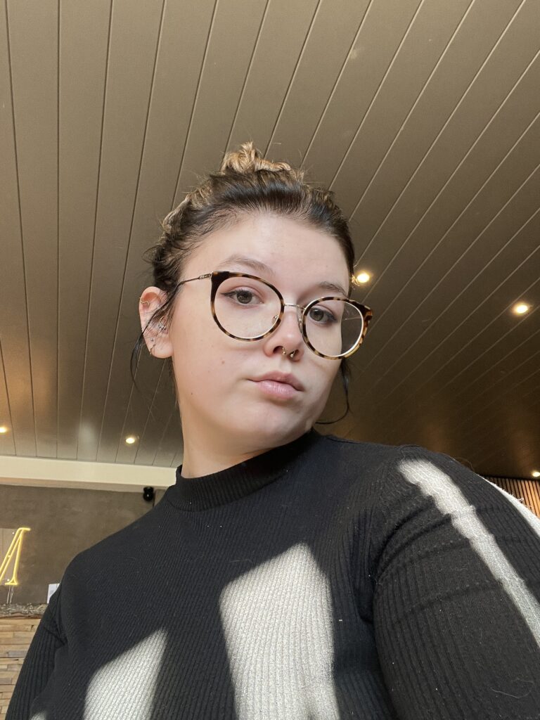 Een portretfoto van tattoo-artiest Phoebe (Tila Bones) op de Sint-Niklaas Tattoo Expo. Ze draagt een bril met montuur in schildpadmotief en een zwarte top, terwijl ze recht in de camera kijkt tegen een achtergrond met sfeervolle verlichting.