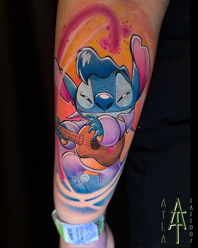 Een kleurrijke tattoo van Stitch (Disney) verkleed als Elvis Presley, spelend op een ukelele. De tattoo knalt van de arm met levendige blauwe, paarse en roze tinten tegen een warme, geel-oranje achtergrond. Gezet door Ayla van Ayla Tattoos op de Sint-Niklaas Tattoo Expo.