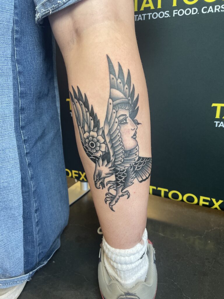 Een gedetailleerde black and grey tattoo op een onderbeen in traditionele stijl. De afbeelding toont een krachtige adelaar met gespreide vleugels, waarbij het gezicht van een vrouw subtiel is verwerkt in een van de vleugels. Gezet door Jordy van Jordy Donders Tattoo op de Sint-Niklaas Tattoo Expo.