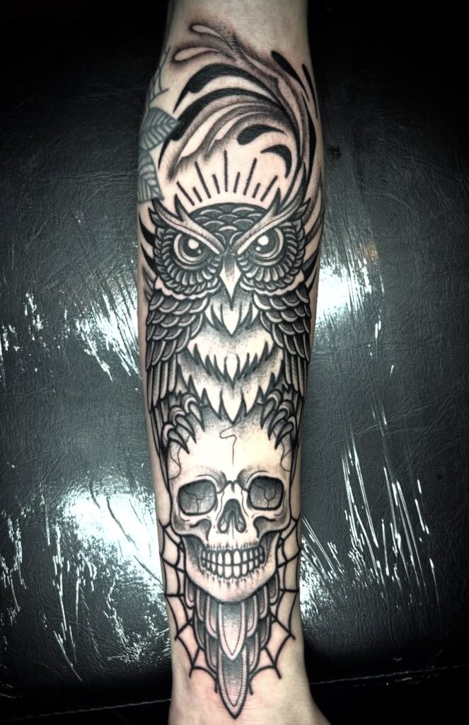 Een krachtige blackwork tattoo op een onderarm van een uil met doordringende ogen die bovenop een menselijke schedel zit. De compositie is verfijnd met grafische lijnen, een spinnenweb aan de onderkant en een stralende zon achter de kop van de uil. Gezet door Daria van Blue Eyes Tattoo op de Sint-Niklaas Tattoo Expo.
