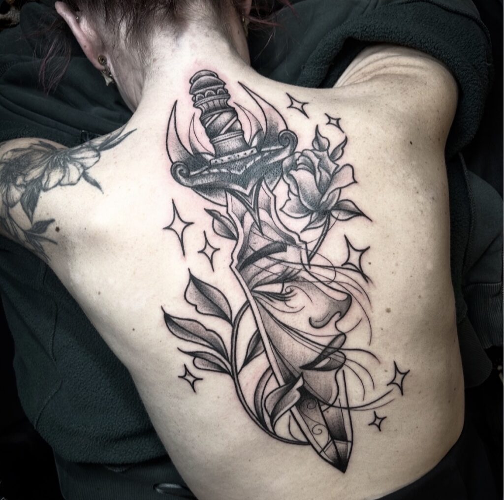 Een indrukwekkende rugtattoo van een rijk versierde dolk die verticaal over de ruggengraat loopt. De dolk snijdt door een vrouwelijk gezicht dat in scherven uiteenvalt, omringd door bloemen, bladeren en fonkelende sterren. Een meesterwerk in zwart-wit door Daria (Blue Eyes Tattoo) op de Sint-Niklaas Tattoo Expo.