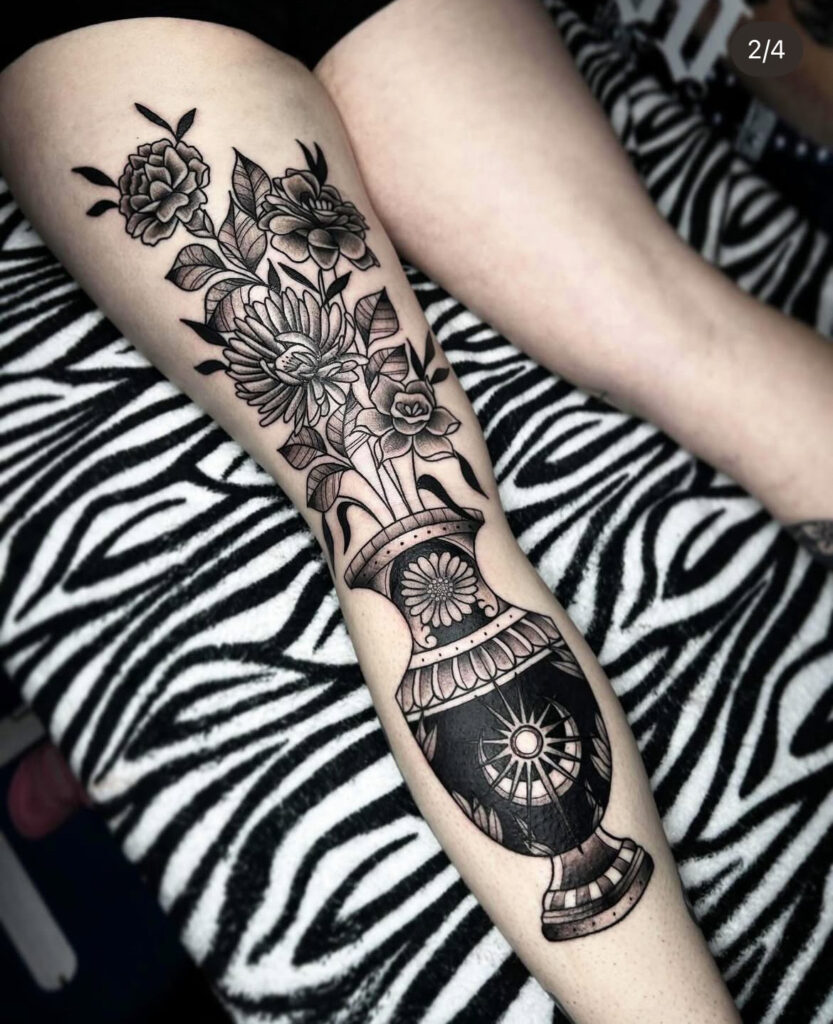 Een grootschalige tattoo op een scheenbeen van een klassieke zwarte vaas met een zon- en maanmotief. Uit de vaas komt een rijk boeket van diverse bloemen zoals pioenrozen en chrysanten, uitgevoerd met diepe zwarttinten en fijn schaduwwerk. Werk van Daria (Blue Eyes Tattoo) op de Sint-Niklaas Tattoo Expo.
