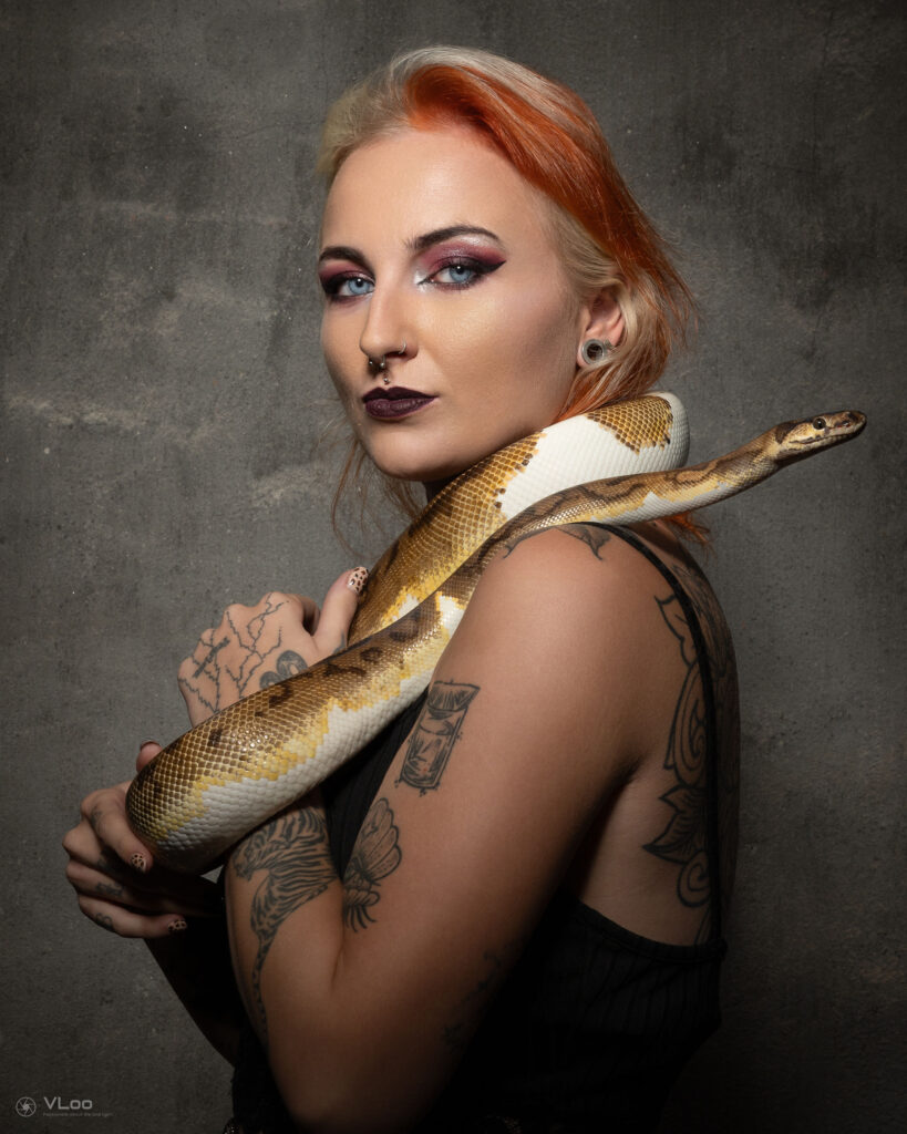 Een krachtig studio-portret van tattoo-artiest Daria (Blue Eyes Tattoo) tegen een grijze betonnen muur. Ze heeft opvallend tweekleurig haar (blond en oranje), diverse gezichtspiercings en draagt een slang over haar schouders. Haar eigen armtattoos en intense blik versterken de artistieke uitstraling.