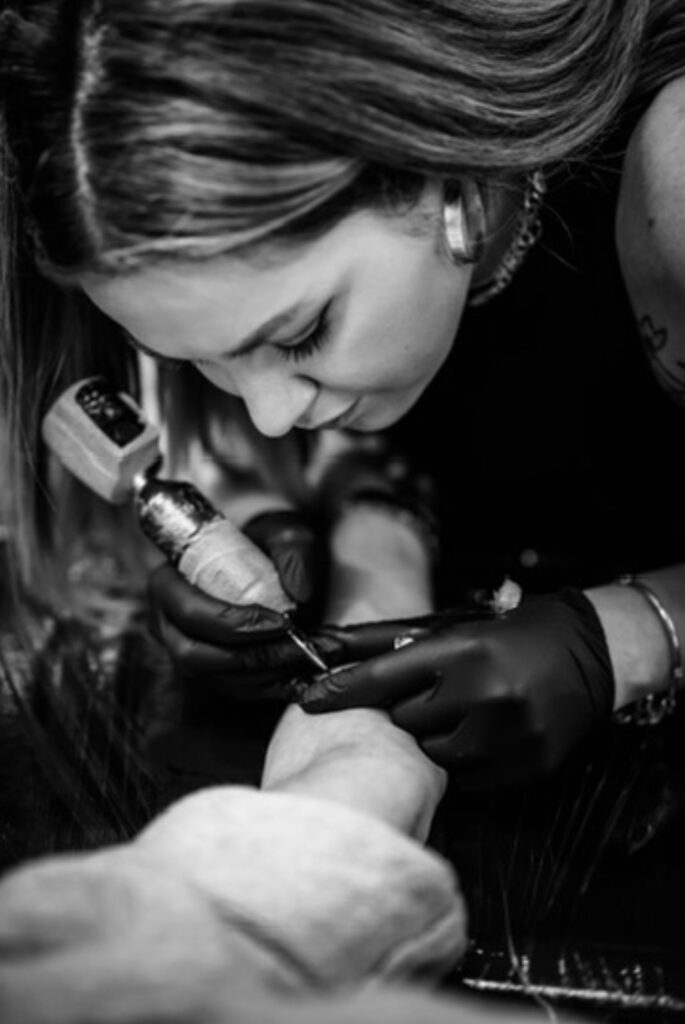 Een sfeervolle zwart-wit foto van tattoo-artiest Sasha (Sasha Hemelaer Tattoo) die geconcentreerd aan een tattoo werkt tijdens de Sint-Niklaas Tattoo Expo. Ze draagt zwarte handschoenen en een professionele tattoo-machine, terwijl haar focus op het fijne lijnwerk duidelijk zichtbaar is.