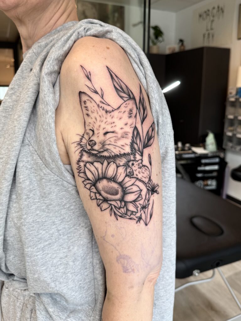 Een hartverwarmende illustratieve tattoo op een bovenarm van een slapende vos, een kleine muis en een zonnebloem. De compositie is verfijnd met botanische elementen en zachte dotwork-schaduwen. Werk van Morgan (Morgan le Fay) tijdens de Sint-Niklaas Tattoo Expo.