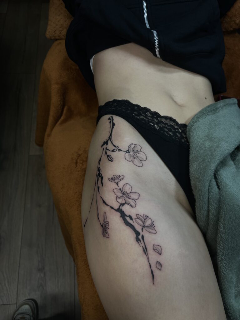 Een elegante en minimalistische tattoo van een kersenbloesemtak op de heup en het bovenbeen. De tak is uitgevoerd met krachtige zwarte lijnen en fijn gearceerde bloemen met vallende blaadjes. Gezet door Amber van Vortex Tattoo tijdens de Sint-Niklaas Tattoo Expo.