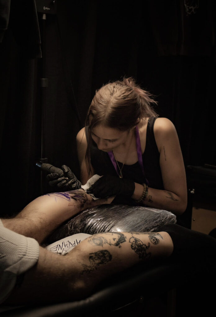 Een sfeervolle actiefoto van tattoo-artiest Amber (Vortex Tattoo) op de Sint-Niklaas Tattoo Expo. Ze is diep geconcentreerd bezig met het zetten van een tattoo op een been, terwijl haar eigen handtattoos en artistieke focus de toewijding aan haar vak tonen.
