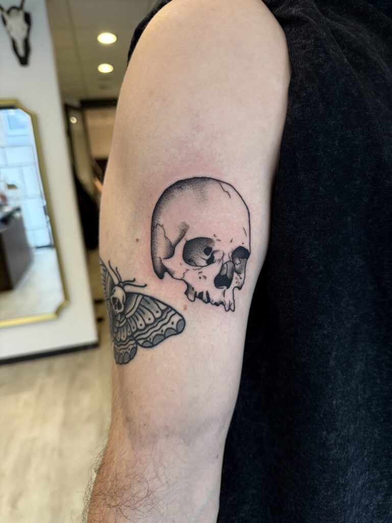 Een illustratieve black and grey tattoo van een menselijke schedel op een bovenarm, gecombineerd met een nachtvlinder (death's-head hawkmoth) aan de zijkant. De stijl kenmerkt zich door strakke lijnen en zacht schaduwwerk. Gezet door Morgan van Morgan le Fay op de Sint-Niklaas Tattoo Expo.