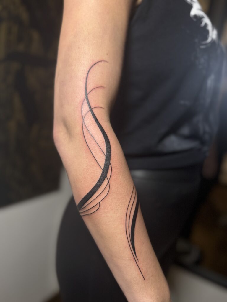 Een minimalistische maar krachtige fine-line tattoo op een onderarm, bestaande uit vloeiende, abstracte zwarte lijnen die de natuurlijke vormen van het lichaam volgen. Een subtiel staaltje vakmanschap van Brett (Tattoossenon) op de Sint-Niklaas Tattoo Expo.