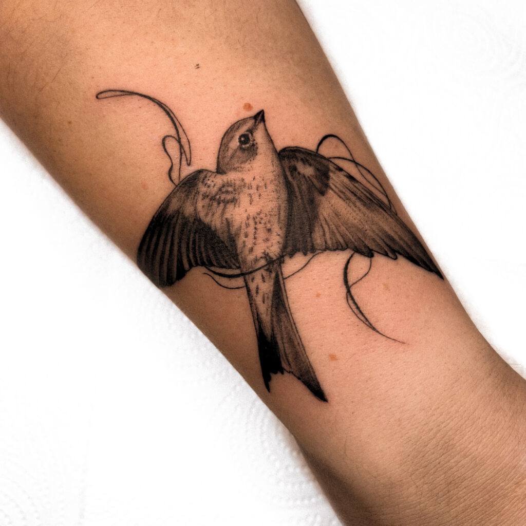 Een gedetailleerde black and grey tattoo van een vogel in volle vlucht op een arm. De vogel is omringd door subtiele, golvende lijnen die beweging suggereren. De focus ligt op de textuur van de veren en de realistische blik van de vogel. Gezet door Chelsy van L'amour Noir Tattoo op de Sint-Niklaas Tattoo Expo.
