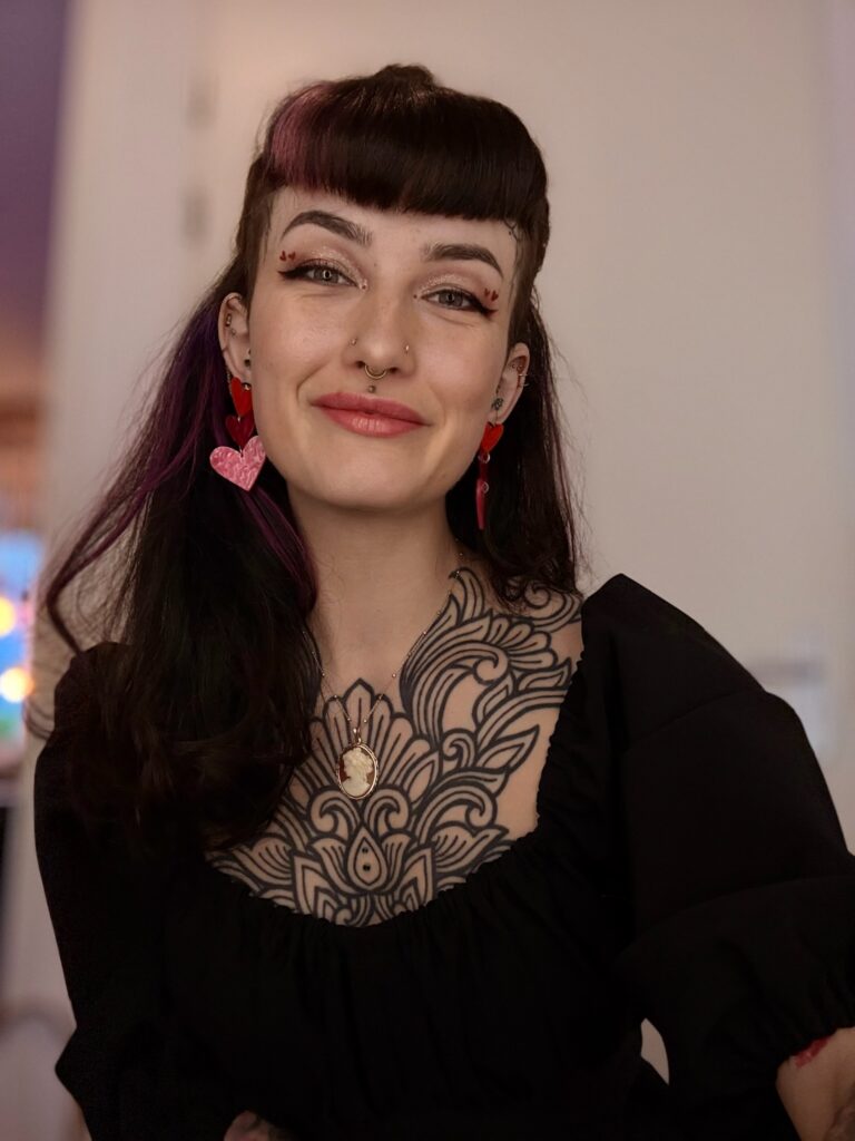 Een portret van tattoo-artiest Ayla (Ayla Tattoos) tijdens de Sint-Niklaas Tattoo Expo. Ze glimlacht vriendelijk naar de camera en draagt een zwarte outfit die haar indrukwekkende mandala-borststuk en een cameo-ketting accentueert. Haar donkere haar heeft paarse accenten en ze draagt opvallende hartvormige oorbellen.