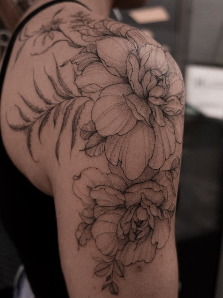 Een grootschalige botanische tattoo op de schouder en bovenarm, bestaande uit pioenrozen gecombineerd met elegante varens. Het ontwerp volgt de natuurlijke welving van de schouder en is gezet met uiterst fijne lijnen. Gezet door Sasha van Sasha Hemelaer Tattoo tijdens de Sint-Niklaas Tattoo Expo.