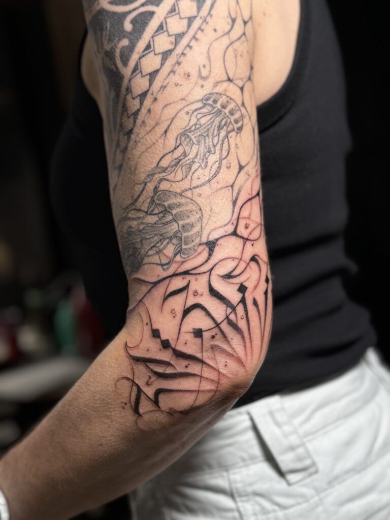 Een gedetailleerde zwart-inkt tattoo op de achterzijde van een arm en elleboog, bestaande uit abstracte calligrafie-elementen (calligraffiti) die vloeiend overgaan in een ontwerp met kwallen. Gezet door Brett van Tattoossenon op de Sint-Niklaas Tattoo Expo.