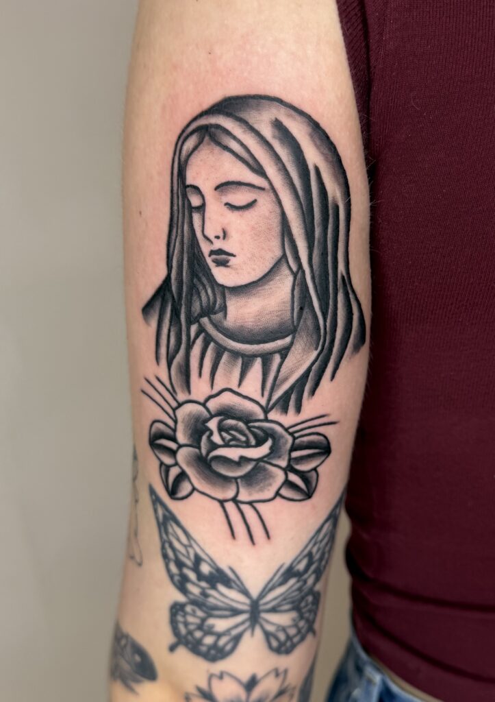 Een verfijnde black and grey tattoo op een bovenarm van de Heilige Maagd Maria met neergeslagen ogen, geplaatst boven een klassieke roos. De tattoo is uitgevoerd in een traditionele stijl met zacht schaduwwerk. Onderaan is een deel van een vlindertattoo zichtbaar. Gezet door Jordy Donders Tattoo tijdens de Sint-Niklaas Tattoo Expo.