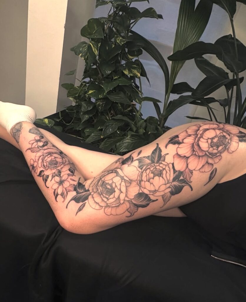 Een grootschalige, elegante tattoo van pioenrozen die over de gehele lengte van het been loopt, van de heup tot de enkel. De bloemen zijn uitgevoerd in een verfijnde black and grey stijl met zachte schaduwen. Gezet door Simon van Sunrise Skin Art op de Sint-Niklaas Tattoo Expo.