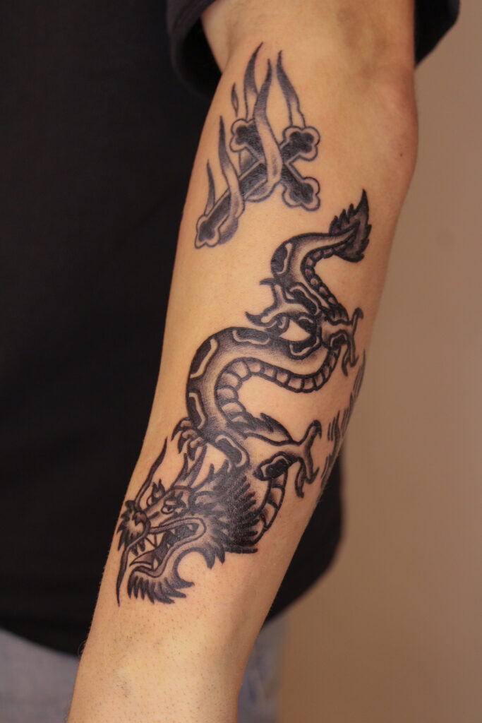 Een strakke blackwork tattoo op een onderarm van een kronkelende Chinese draak. De draak is uitgevoerd met stevige zwarte lijnen en schaduwwerk. Daarboven is een kleiner ontwerp van een vlammend kruis met botten zichtbaar. Werk van Jordy (Jordy Donders Tattoo) op de Sint-Niklaas Tattoo Expo.