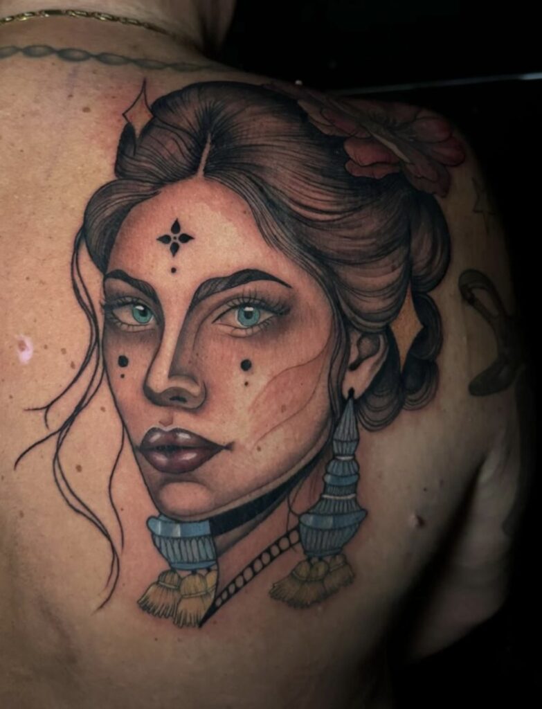 Een prachtig, dromerig portret van een vrouw getatoeëerd op een rug. Het ontwerp bevat zachte kleurovergangen, blauwe ogen en decoratieve elementen zoals een bloem in het haar en opvallende oorbellen. Werk van Sanne (Santji Art) op de Sint-Niklaas Tattoo Expo.
